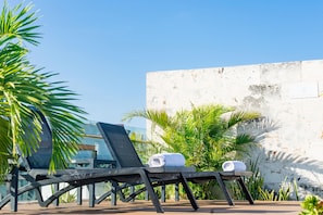 Un área de terraza con sillones y una mesa, rodeada de palmeras y un muro blanco.