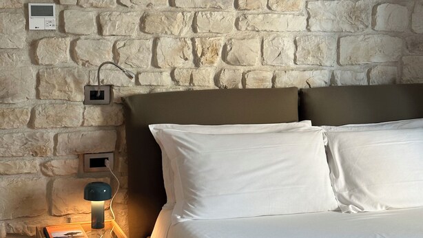 Un dormitorio con pared de piedra, una cama con ropa de cama blanca, una mesita de noche con lámpara y una pequeña mesita auxiliar de madera.