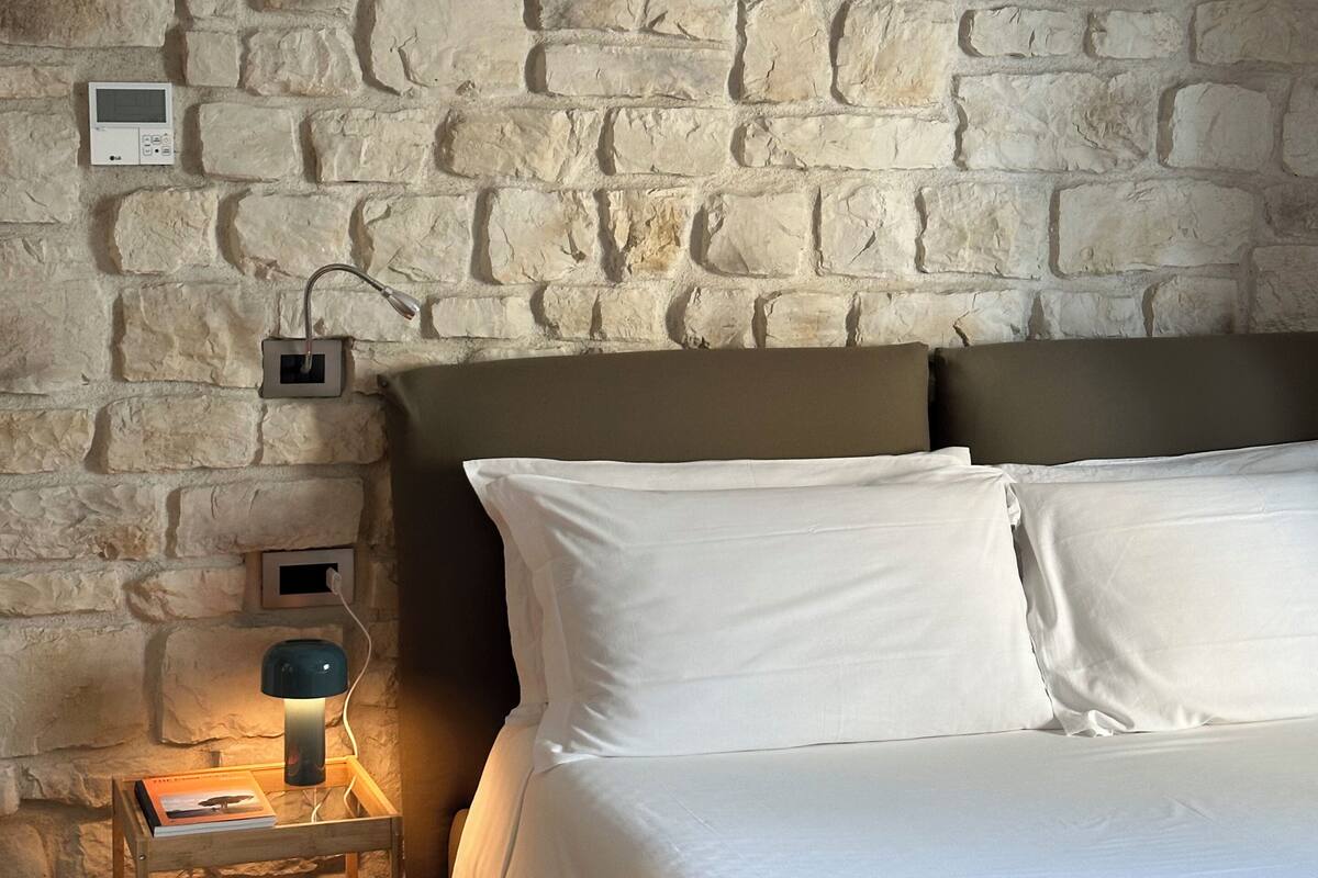 Ein Schlafzimmer mit Steinwand, einem Bett mit weißen Leinen, einem Nachttisch mit Lampe und einem kleinen Holzbeistelltisch.