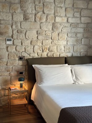 Un dormitorio con pared de piedra, una cama con ropa de cama blanca, una mesita de noche con lámpara y una pequeña mesita auxiliar de madera.