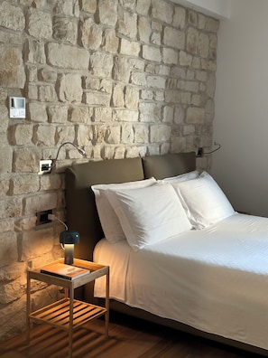 Een slaapkamer met een stenen muur, een bed met witte lakens, een nachtkastje met een lamp en een stopcontact.