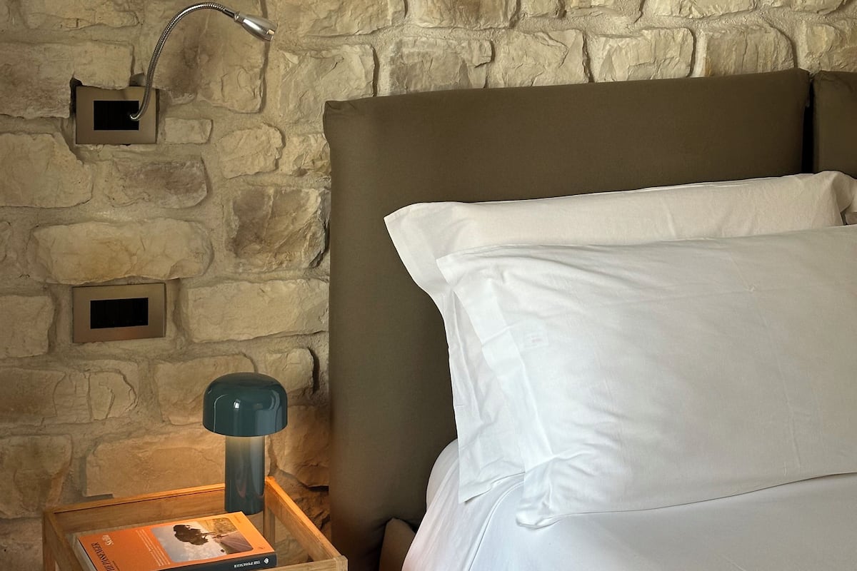 Ein Schlafzimmer mit einer Steinwand, einem hölzernen Nachttisch mit Lampe und einem Bett mit weißen Leinen.