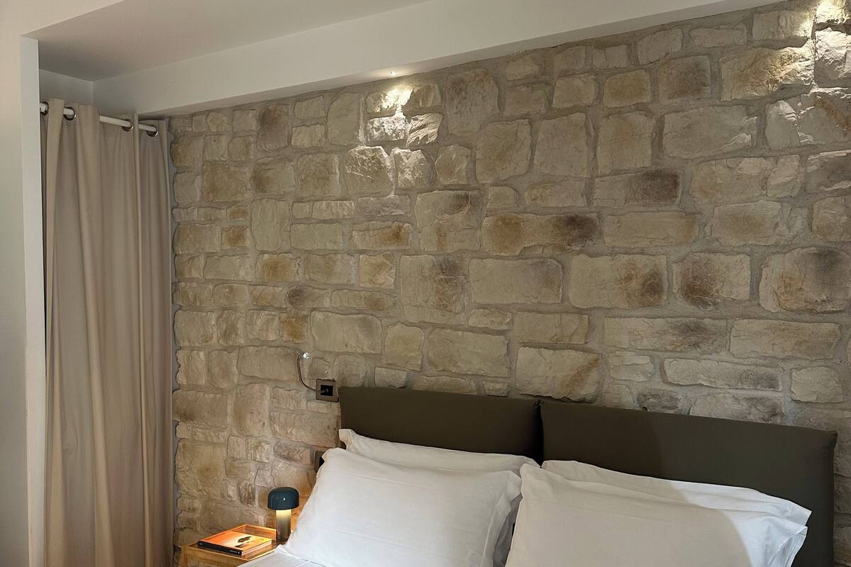Ein Schlafzimmer mit Steinswand, einem Bett mit weißen Bettwäsche und einem Nachttisch mit Lampe.