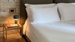Un dormitorio con pared de piedra, una mesita de noche de madera con lámpara y una cama con ropa blanca.