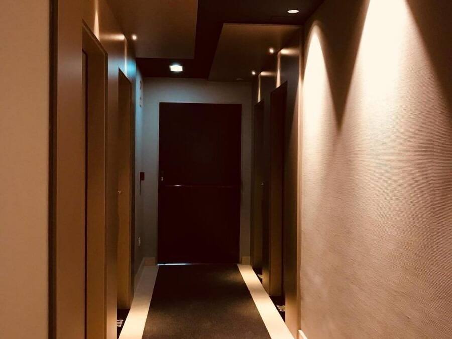 Un estrecho pasillo alfombrado con iluminación empotrada y puertas a ambos lados, que conduce a un cuarto oscuro al final.