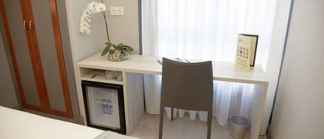 Un dormitorio con una cama, un escritorio con una silla, un refrigerador pequeño, un cesto de basura y una ventana con cortinas.
