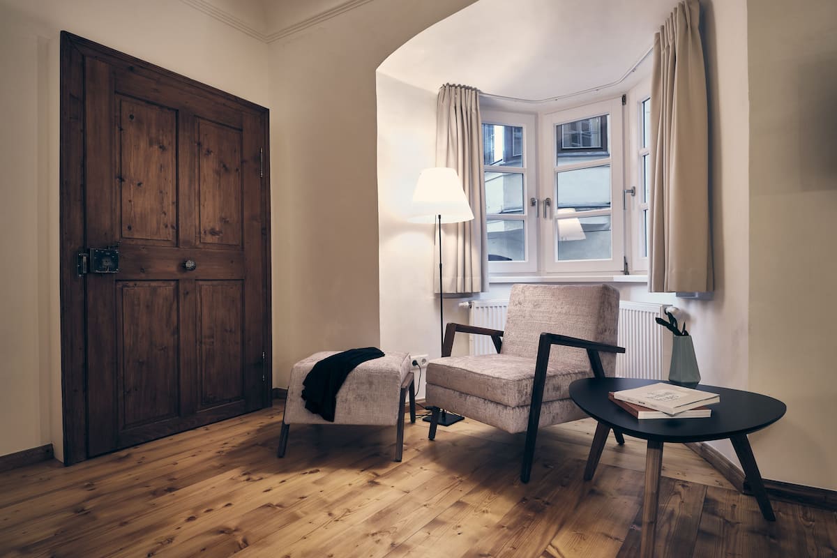 Ein Zimmer mit Holztür, einem fensterbelaubten Fenster, einem Sessel, einem kleinen Tisch mit Lampe und einem Heizkörper.