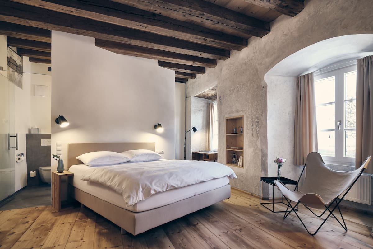Ein Schlafzimmer mit Holzboden, einem Bett mit weißer Bettwäsche, einem Stuhl, einem Schreibtisch und einem Fenster mit Vorhängen.