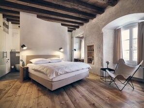 Ein Schlafzimmer mit Holzboden, einem Bett mit weißer Bettwäsche, einem Stuhl, einem Schreibtisch und einem Fenster mit Vorhängen.