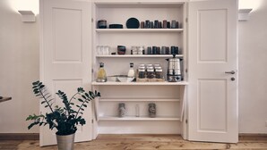 Ein heller Innenraum mit Holzboden, einem Topfpflanzen und einer weißen Schrank mit Regalen, die mit Gläsern, Flaschen und Küchenutensilien gefüllt sind.