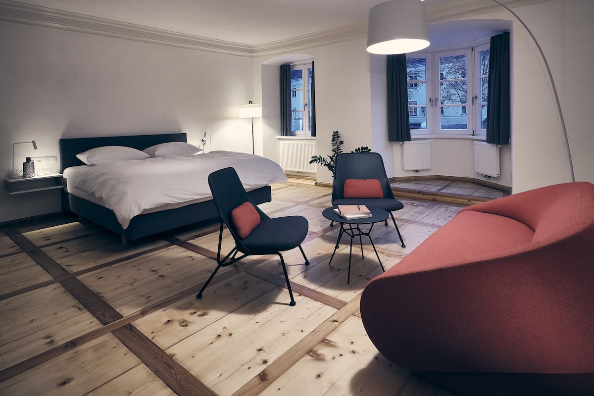Ein modernes Schlafzimmer mit einem großen Bett, zwei Stühlen, einer Stehlampe und Blick ins Freie durch ein Fenster.