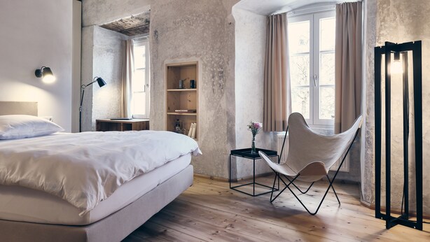 Ein Schlafzimmer mit Holzboden, einem Bett, einem Sessel, einem Regal und einem Fenster mit Vorhängen.