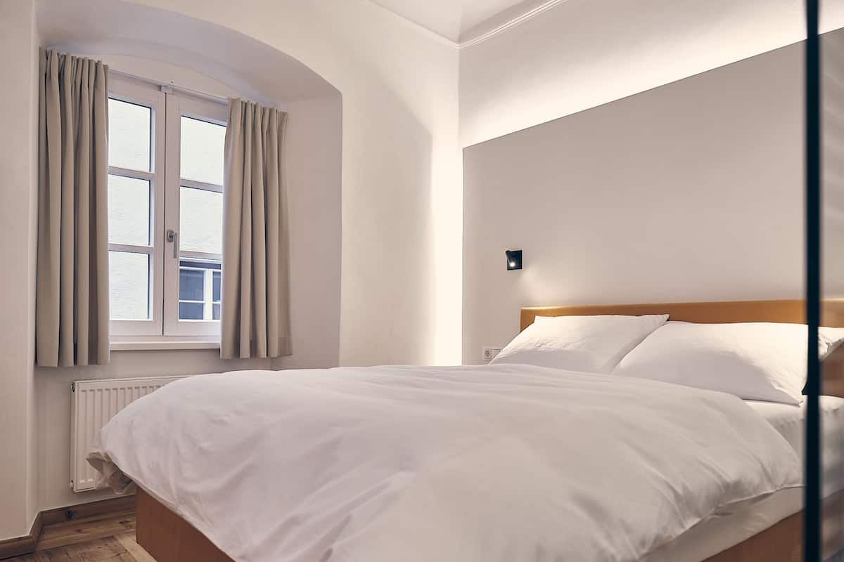 Ein Schlafzimmer mit einem hölzernen Bett, einem Fenster mit Vorhängen, einem Heizkörper und einer Wandleuchte.