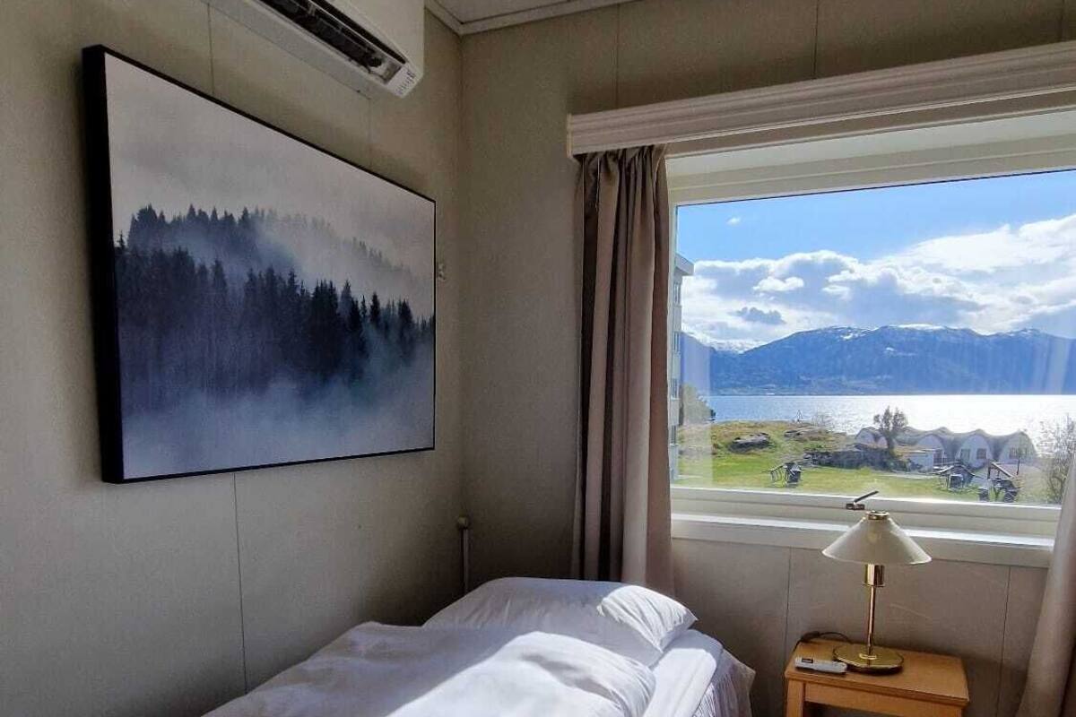 Un dormitorio con una cama individual, un cuadro de un bosque, un equipo de aire acondicionado, una ventana con vistas a un paisaje y una mesita de noche con una lámpara.