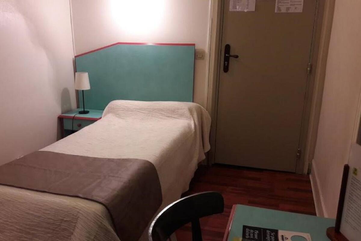 A small, single-bed room with a teal headboard, a bedside table with a lamp, and a door with a lock.