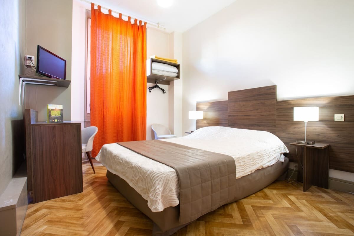 A hotel room with a wooden bed, a desk with a computer, a chair, and a nightstand with a lamp.