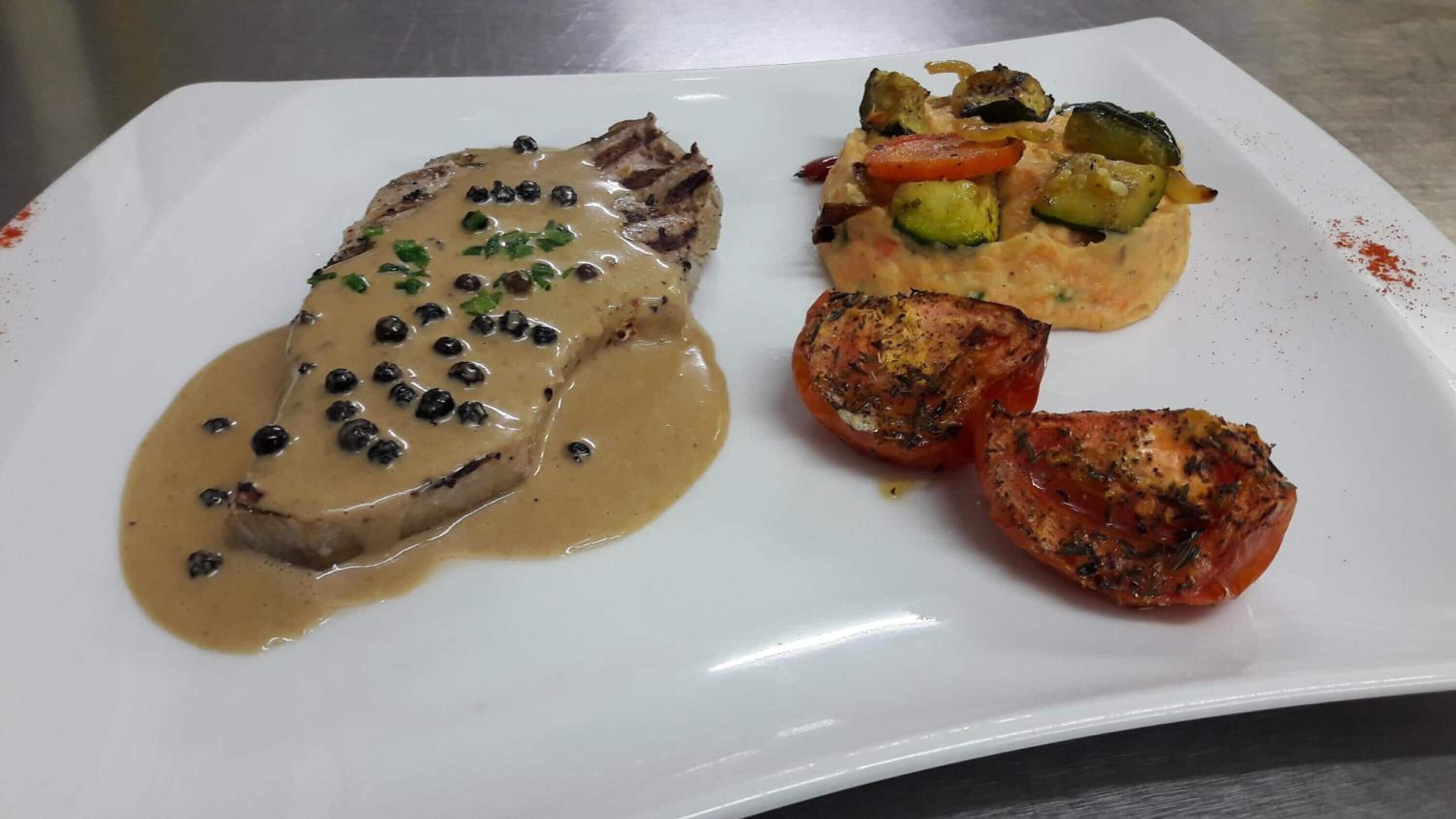 Une assiette contenant un steak nappé d’une sauce crémeuse et parsemé de grains de poivre noir, accompagné de légumes grillés et d’une tomate rôtie.