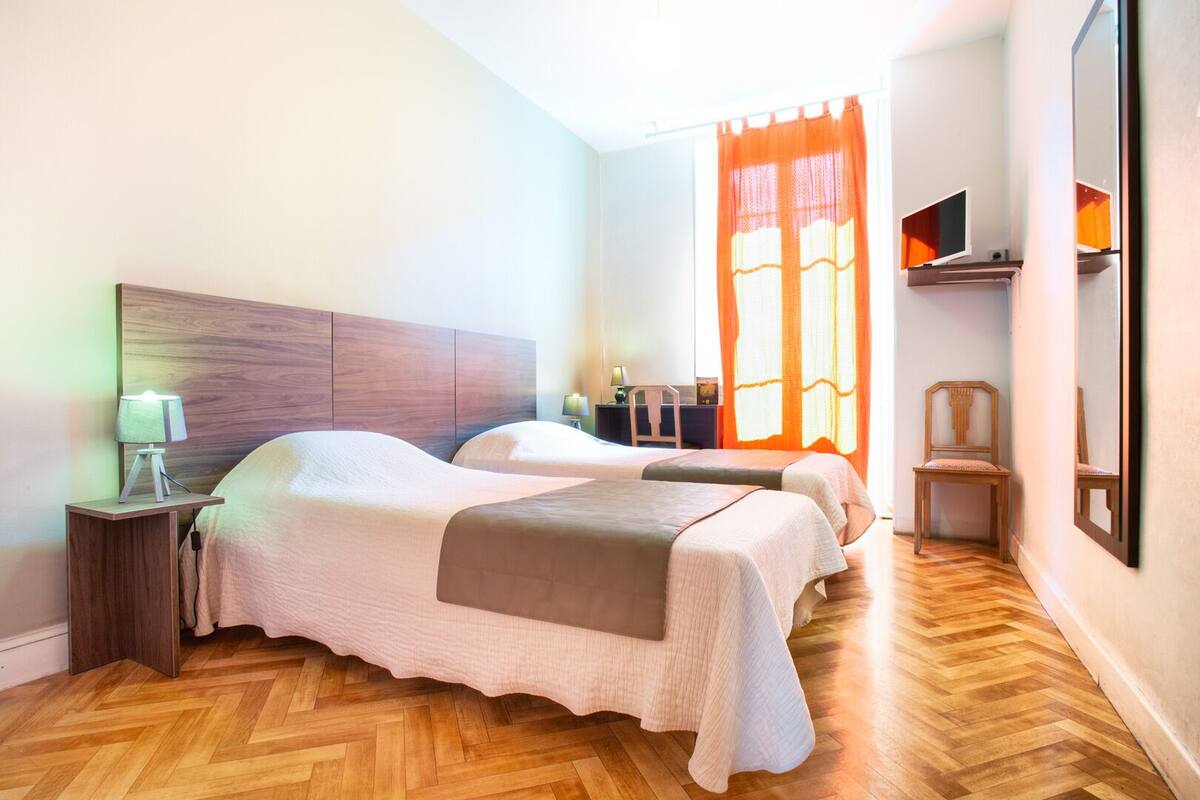 A hotel room with two beds, wooden flooring, a nightstand with a lamp, a chair, and a TV mounted on the wall.