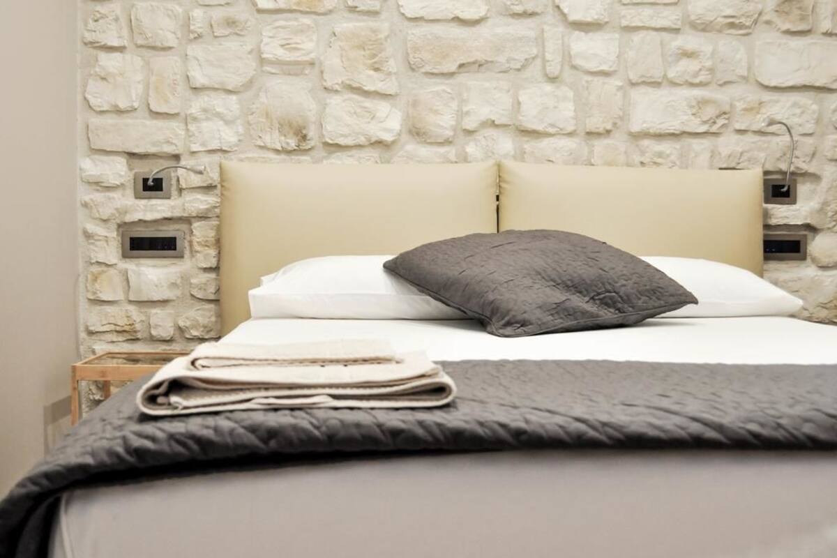 A neatly made bed with a grey blanket and pillows, a wooden nightstand with a folded towel, and a stone wall with electrical outlets.