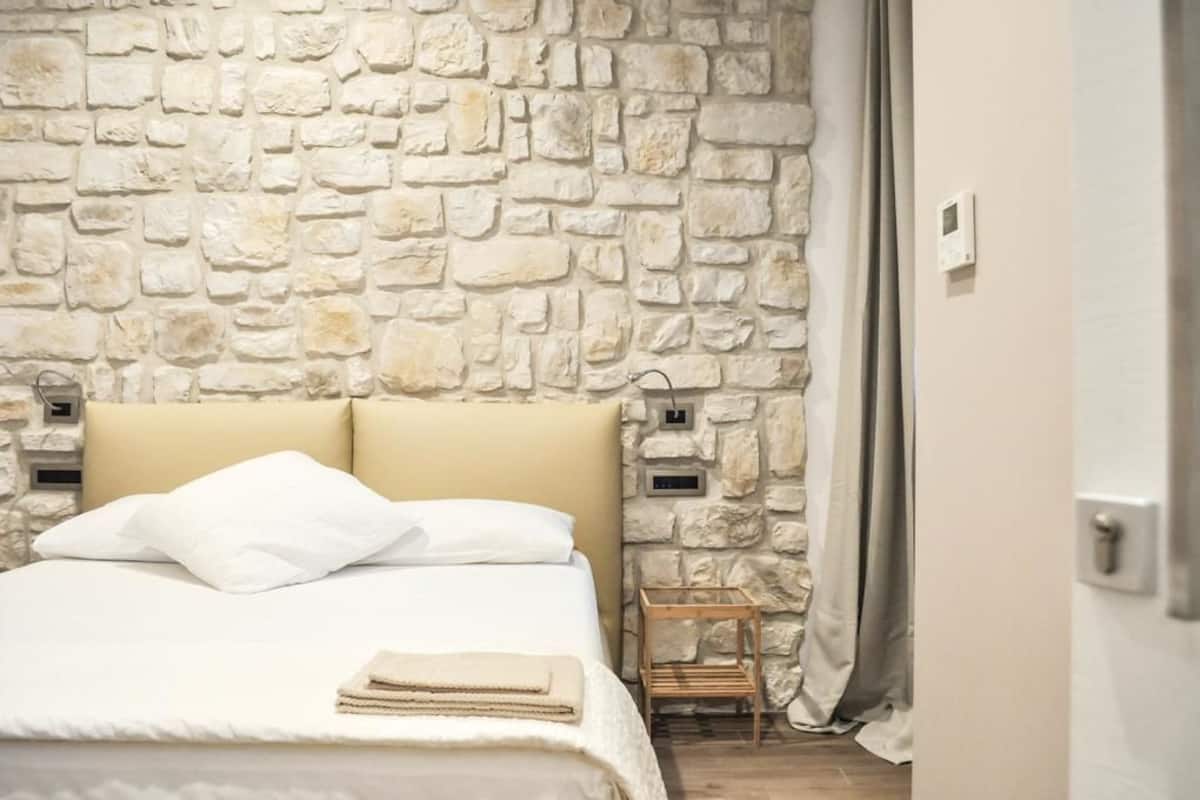 A bedroom with a stone wall, a bed with white linens, a wooden nightstand, and a small table with a lamp.