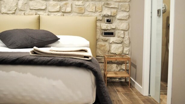 A bedroom with a stone wall, a bed with a dark pillow, a small wooden nightstand, and a door leading to a bathroom. - Olivia Rooms Eurialo (Syracuse)