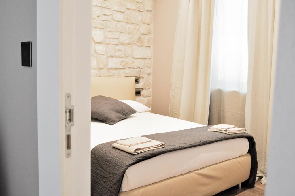 A bedroom with a stone wall, a bed with a beige comforter, a dark blanket, and a folded towel on the bed.