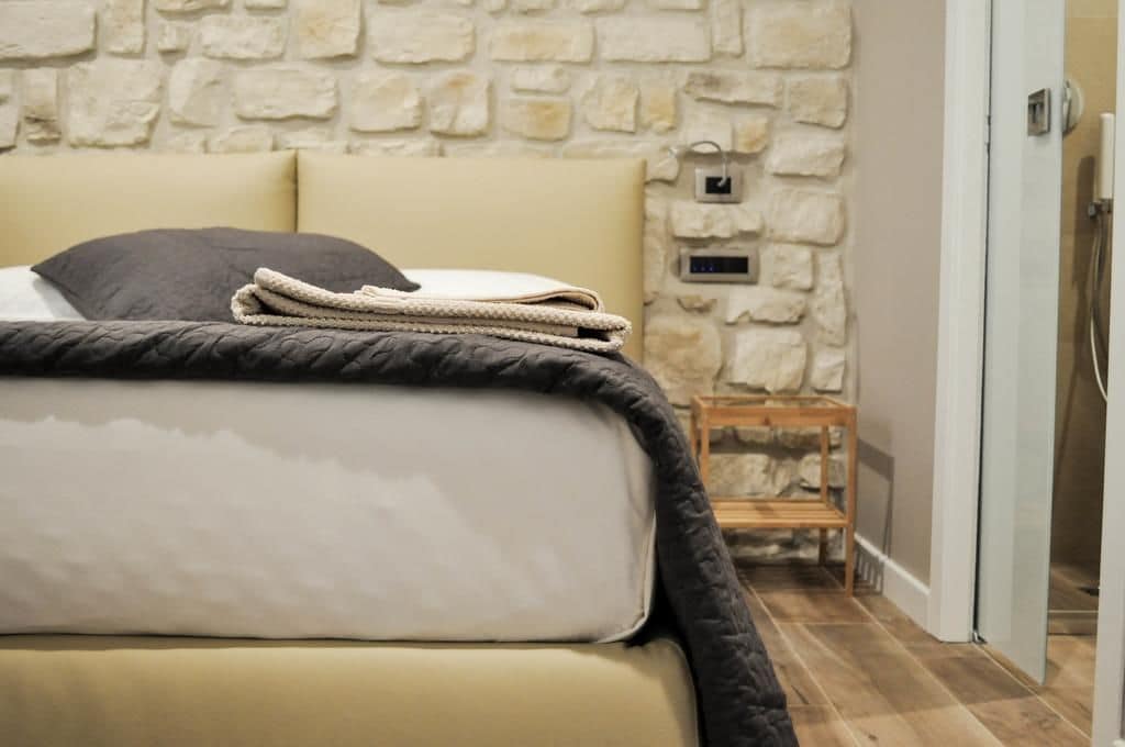 Un lit avec une couverture matelassée et un oreiller, un mur en pierre, une petite étagère en bois et une porte donnant sur une douche.