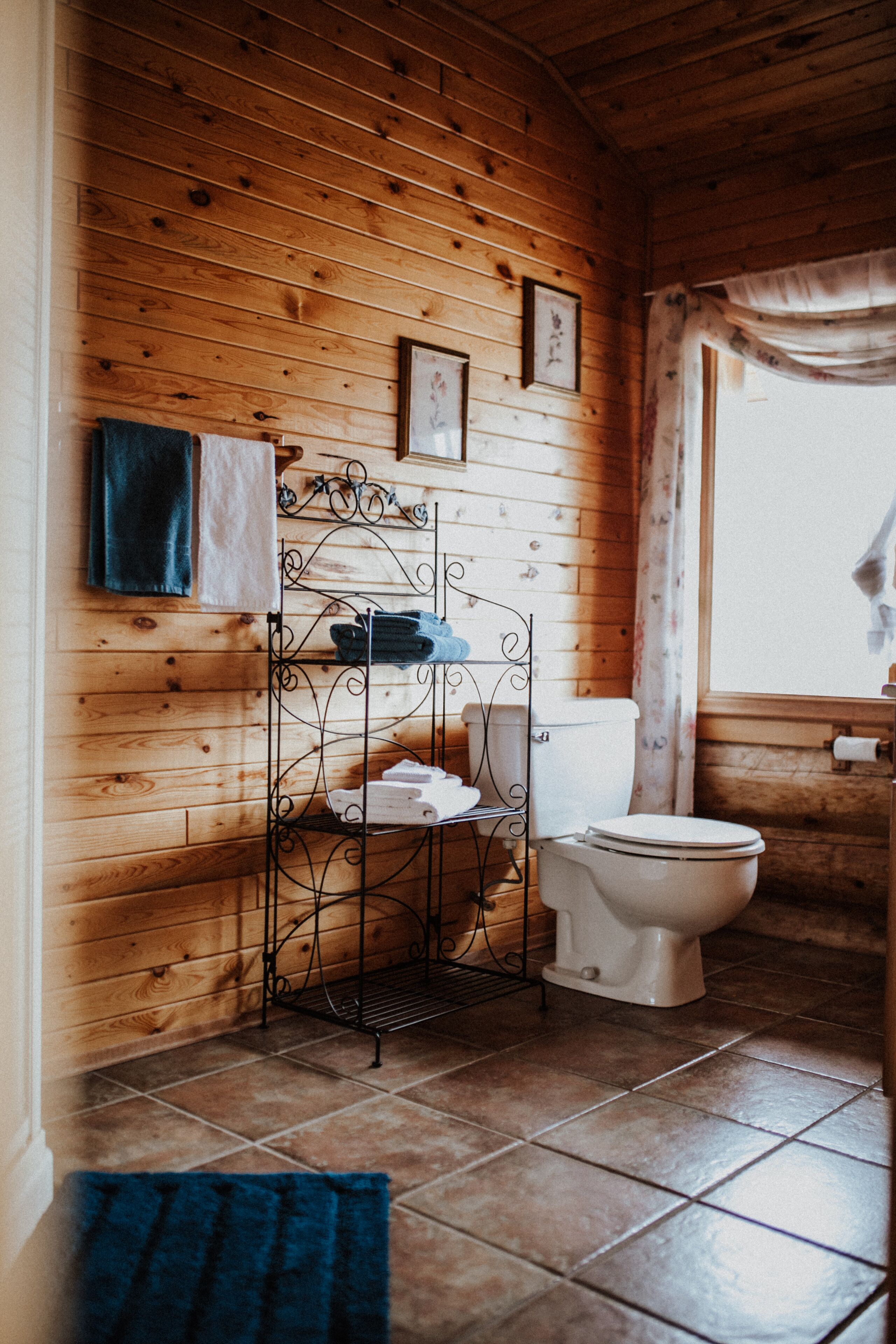 Un baño con paneles de madera, un inodoro, un estante de metal con toallas y una ventana con cortinas.