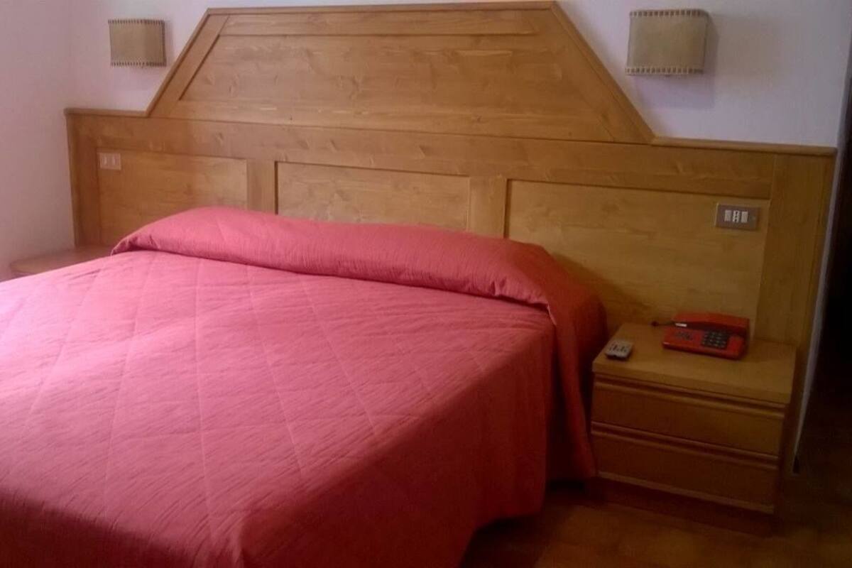 Een kamer met een houten hoofdbord, een bed met een rode spreideken, een nachtkastje met een telefoon en een wandlamp.