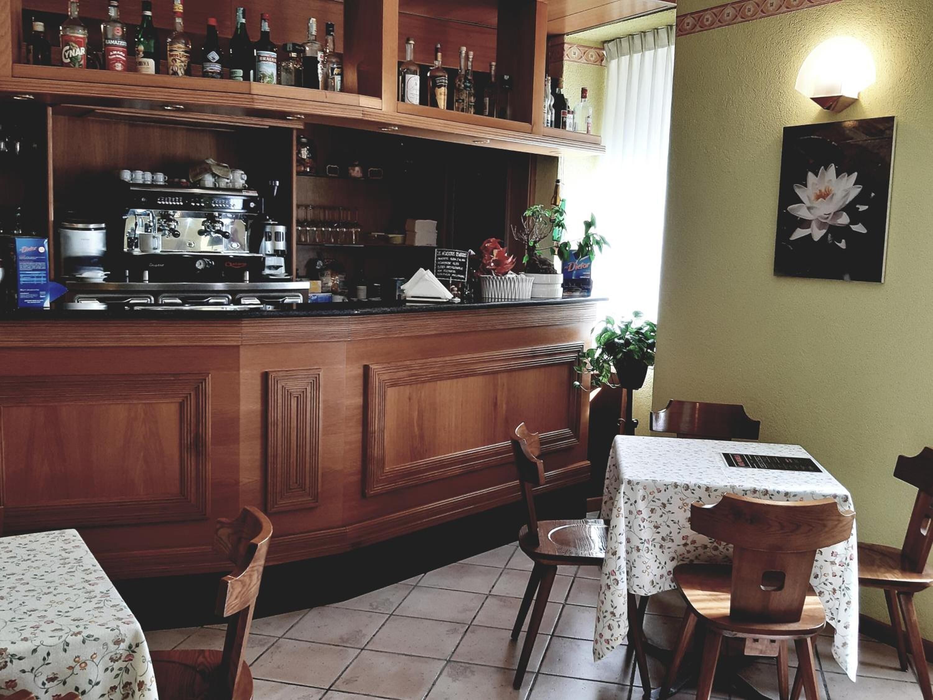 Eine Bar mit Holzwänden, einer Kaffeemaschine und verschiedenen Flaschen. Es gibt Stühle und Tische mit Blumenmuster-Tischdecken.