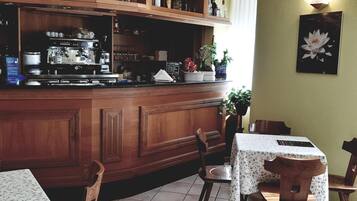 Eine Bar mit Holzwänden, einer Kaffeemaschine und verschiedenen Flaschen. Es gibt Stühle und Tische mit Blumenmuster-Tischdecken.