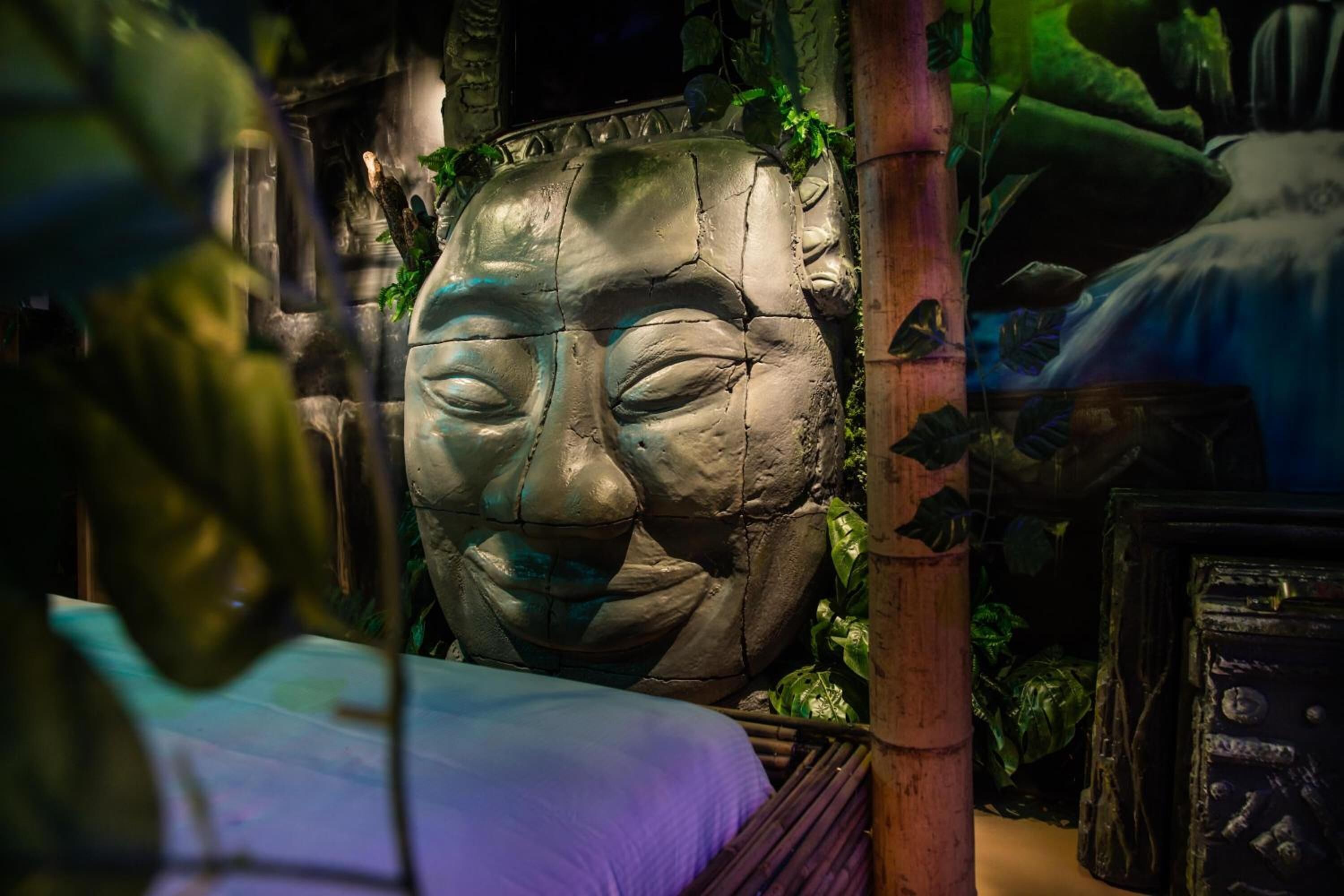 A room with a stone face sculpture, wooden pillars, and a bed with a purple blanket.