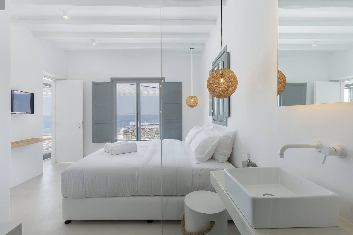 Une chambre moderne avec un grand lit, un lavabo blanc et une vue sur la ville par la fenêtre.