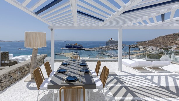 A dining area with a table set for six, overlooking a harbor with boats and a ship. - Sealine Villas (Mykonos)