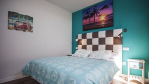 A bedroom with a bed, a checkered headboard, a teal wall, a beach-themed artwork, and a small bedside table. - Apartamentos El Volante II (Ciempozuelos)