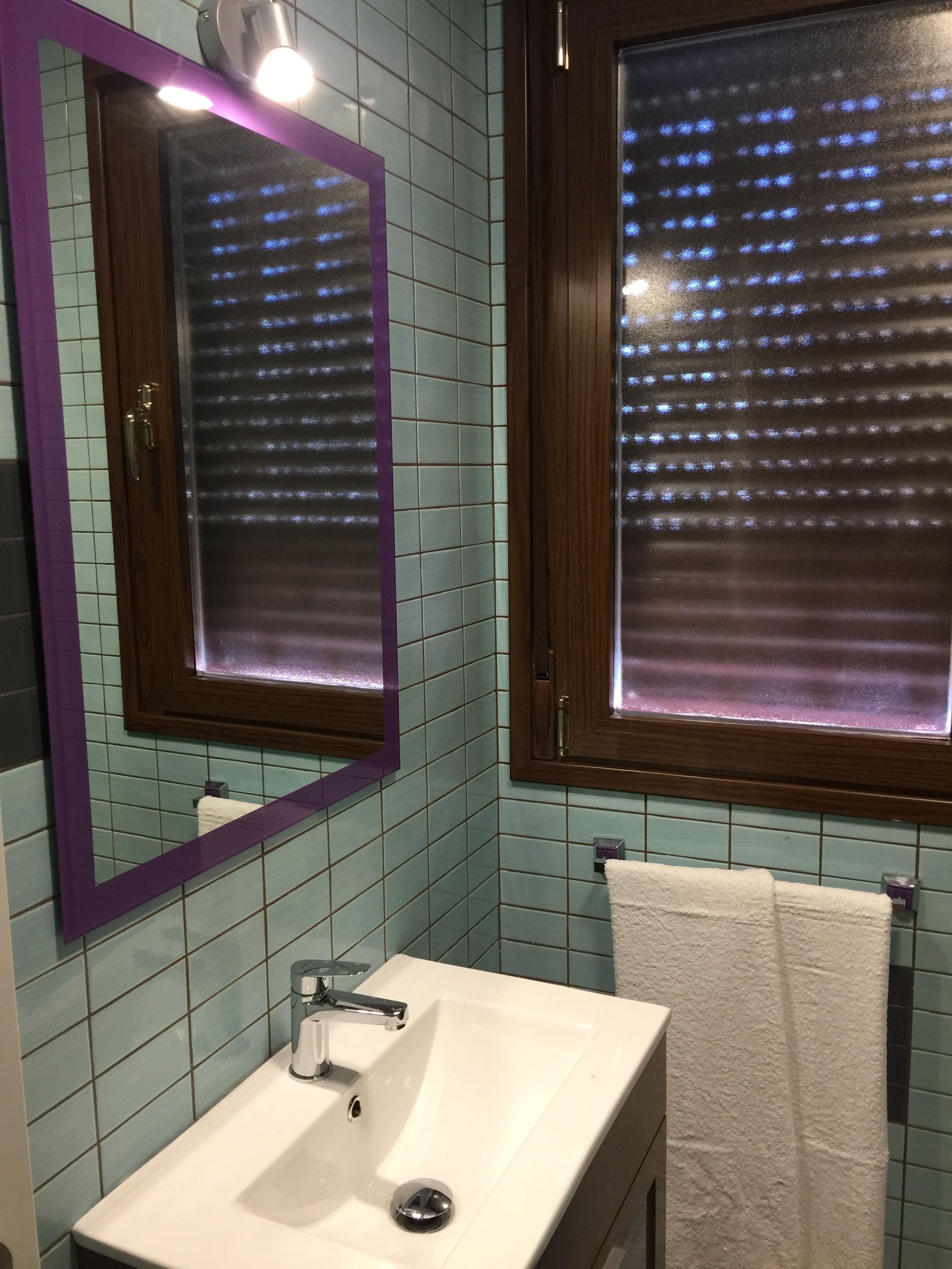 Un baño con lavamanos blanco, un espejo con marco morado y una ventana con persianas.