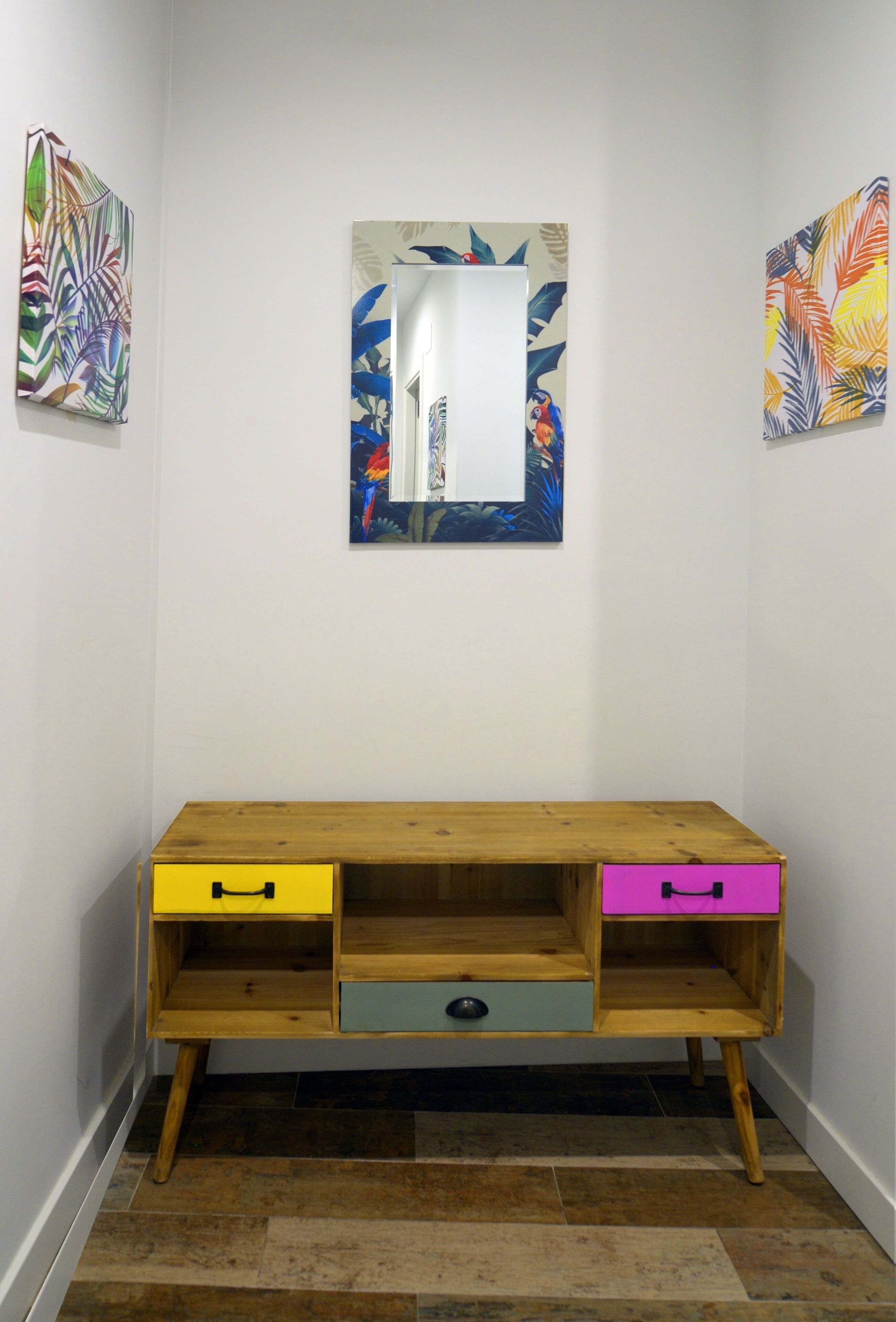 Een houten console tafel met twee lades, één geel en één roze, tegen een witte muur met twee kleurrijke schilderijen met bladmotieven.