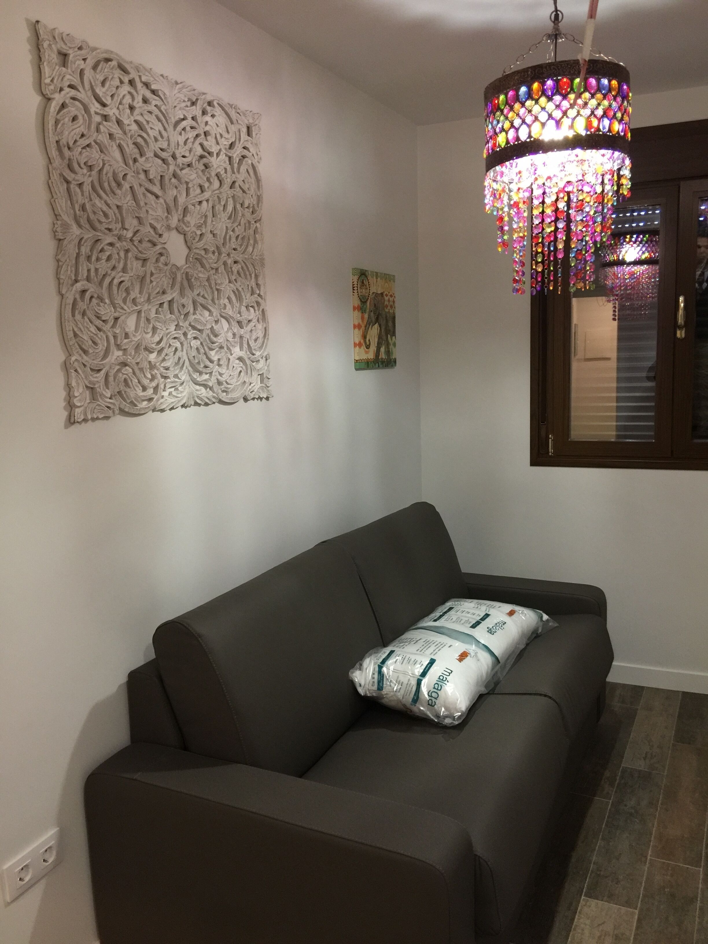 Un salon avec un canapé marron, un élément décoratif mural, un luminaire suspendu aux couleurs vives et une fenêtre avec des stores.