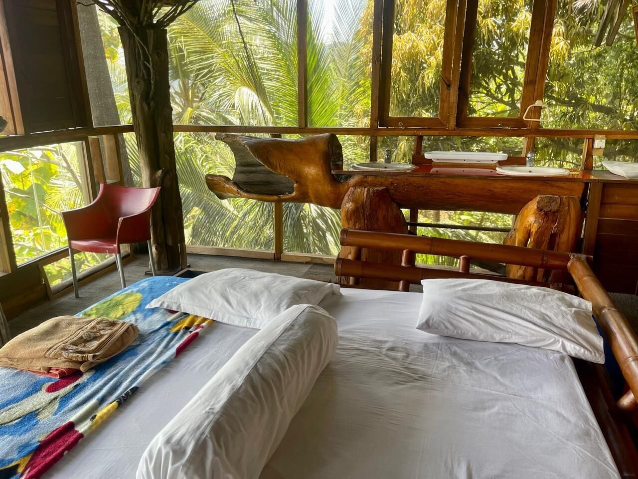 A room with a bed, a wooden headboard, a red chair, and a view of palm trees through large windows.
