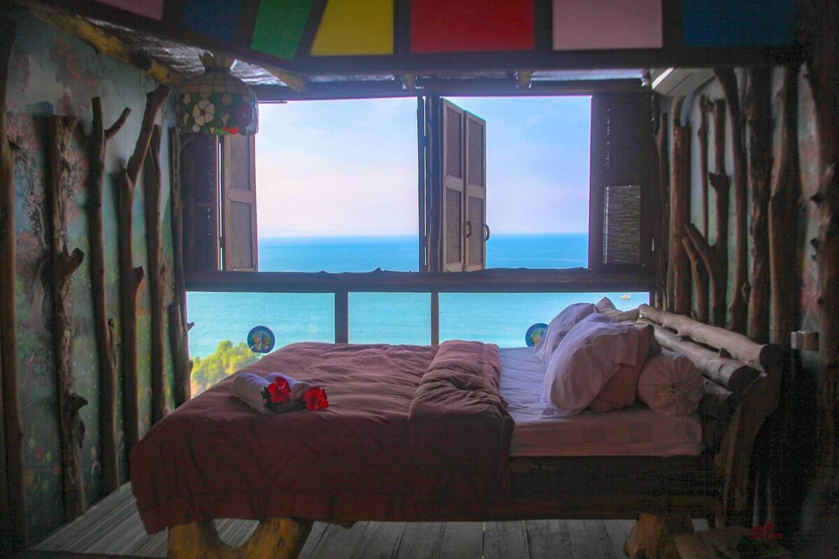 Una cama con vista al océano, piso de madera y ventana con vistas.