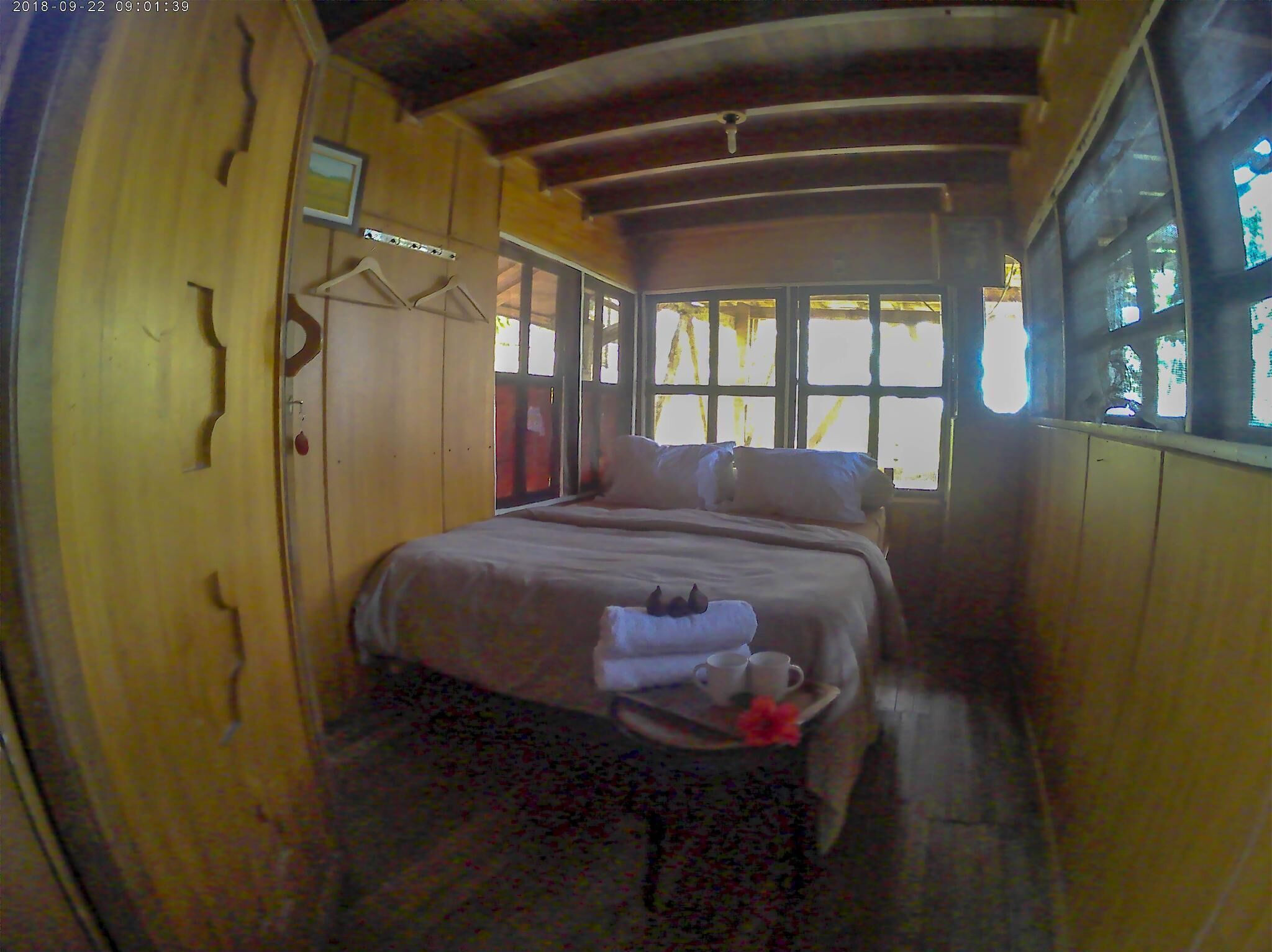 A narrow, wooden room with a bed, a small table with cups and a towel, and a window with natural light.
