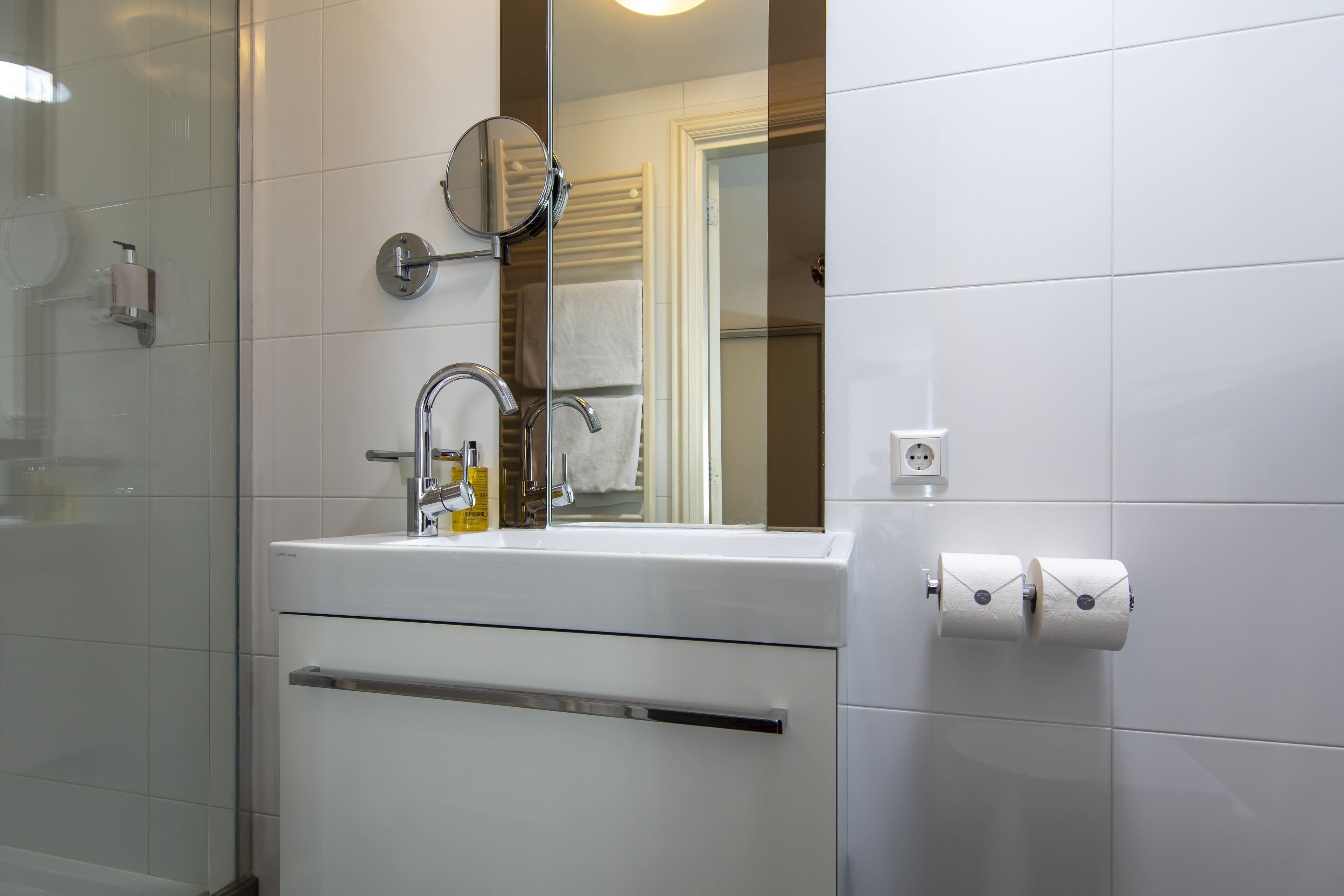 A bathroom with a glass shower enclosure, a white sink with a mirror, and two rolls of toilet paper on the wall.