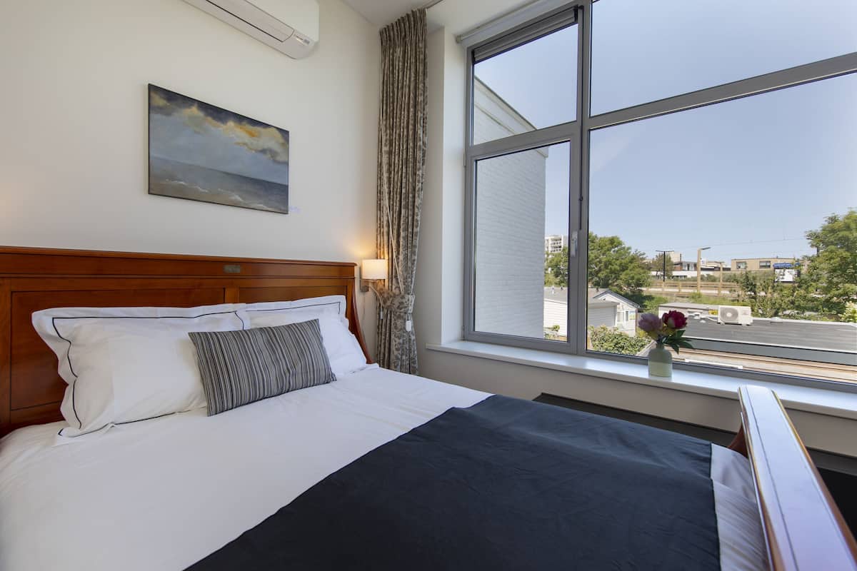 A hotel room with a bed, a window with a view of buildings, a painting on the wall, and a vase with flowers on the windowsill.