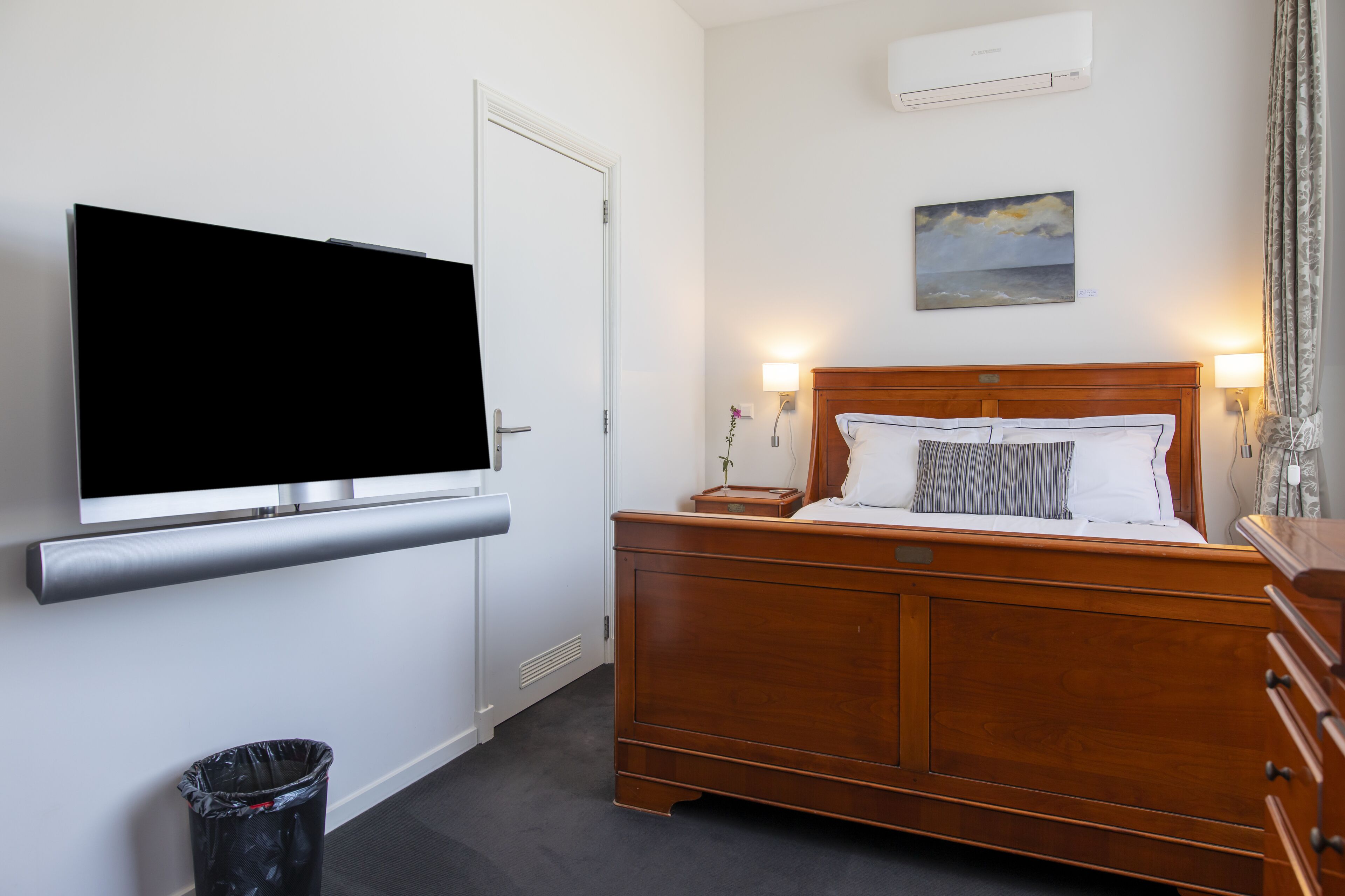 Habitación de hotel con una cama de madera, un televisor de pantalla plana en la pared, una mesita de noche pequeña y un aire acondicionado instalado en la pared.