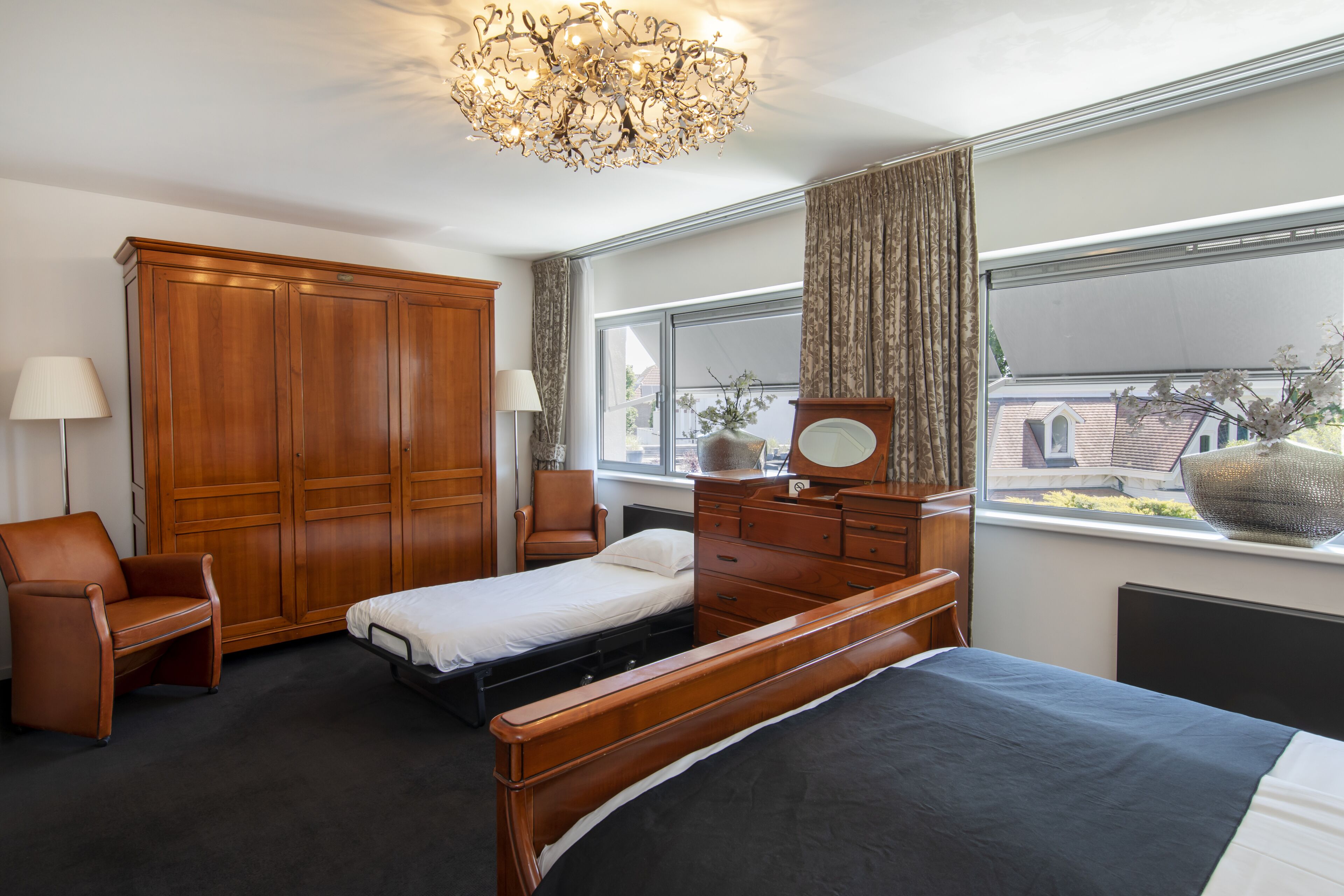 Ein Schlafzimmer mit Bett, Kleiderschrank, Stuhl, Kommode, Spiegel und Fenster mit Ausblick.