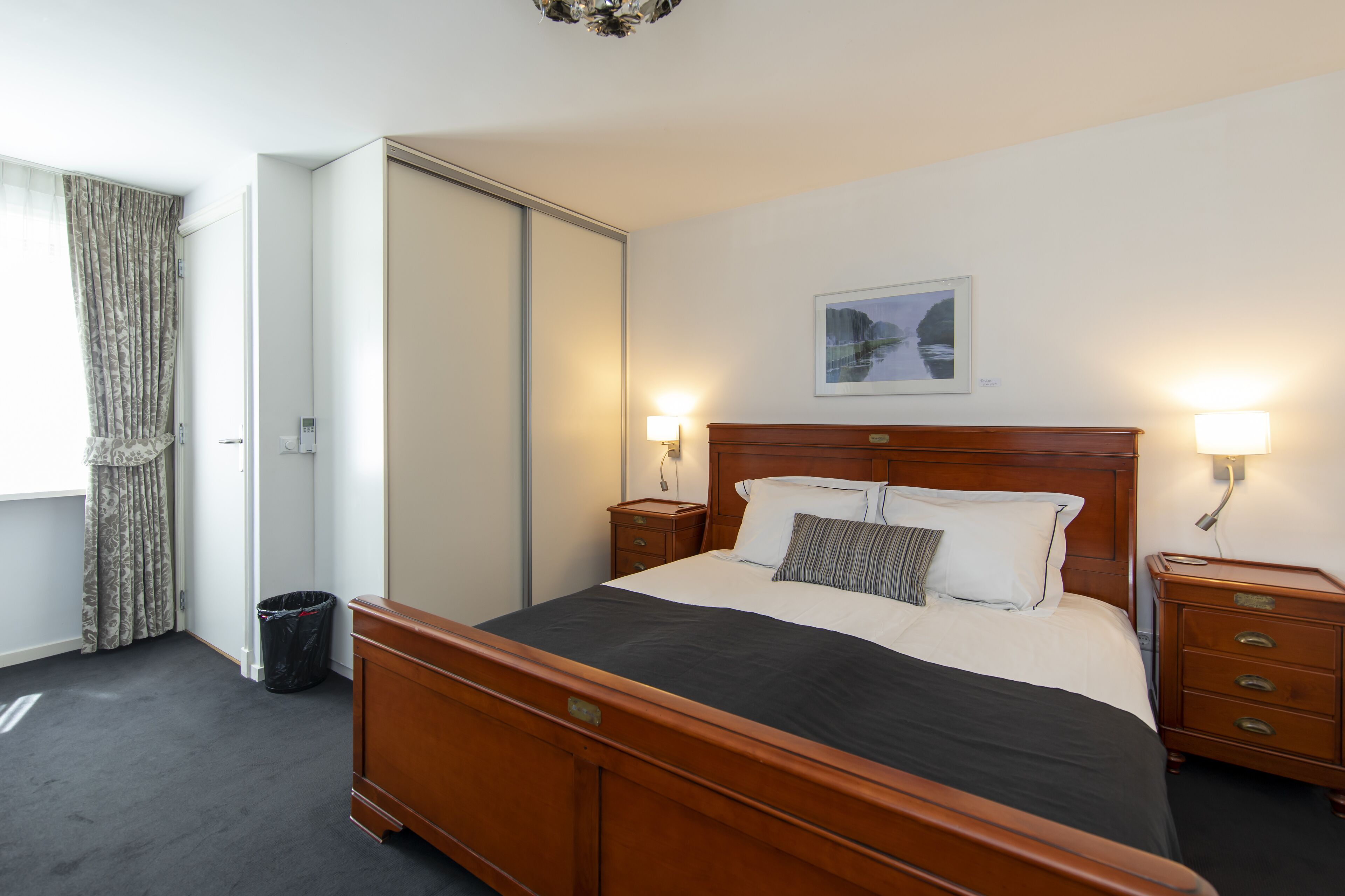 A neatly arranged bedroom with a large bed, bedside tables, a wooden headboard, a framed picture on the wall, and a window with curtains.