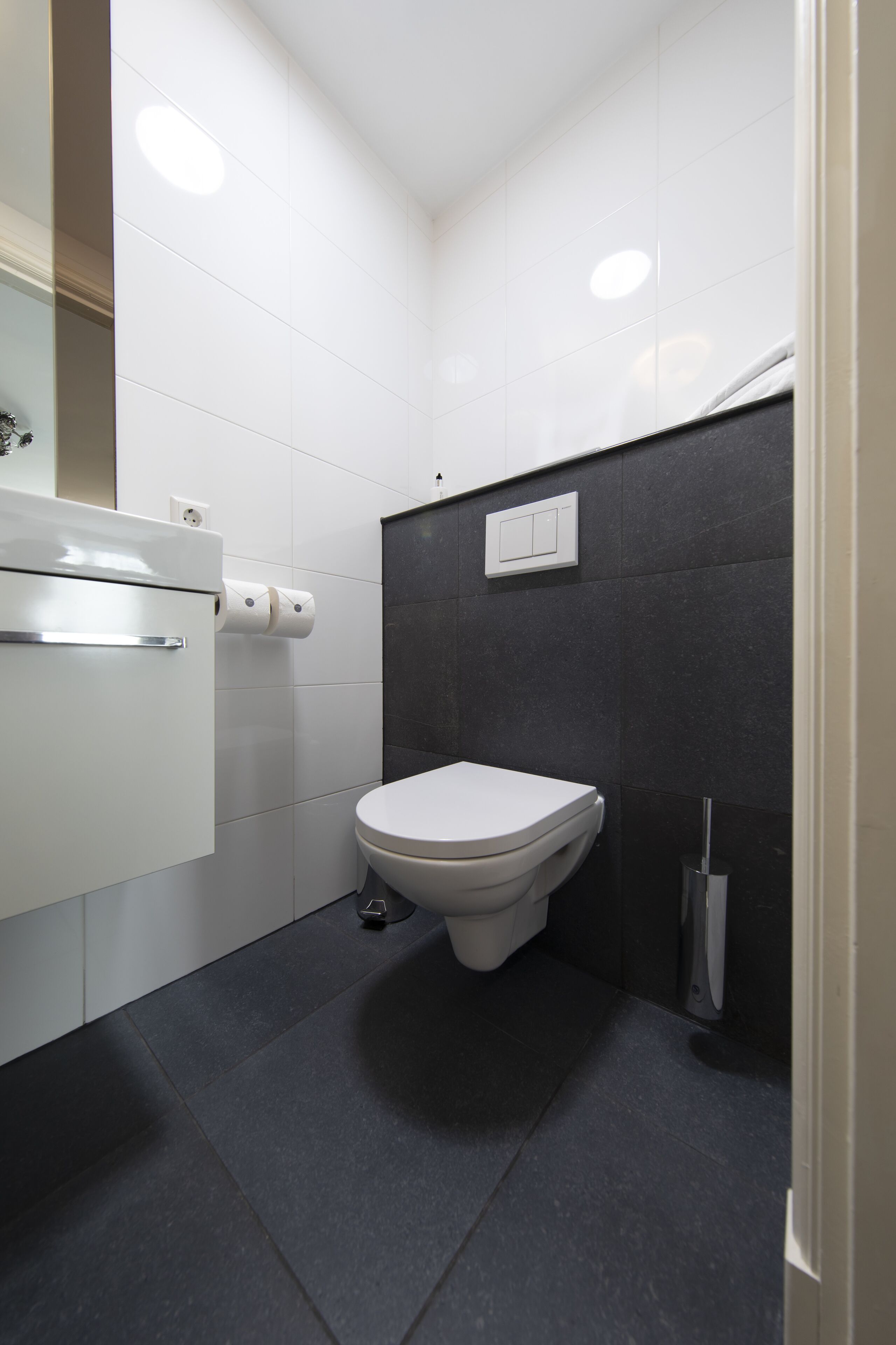 A modern bathroom with a white toilet, dark tiled walls, a white cabinet, and a mirror.