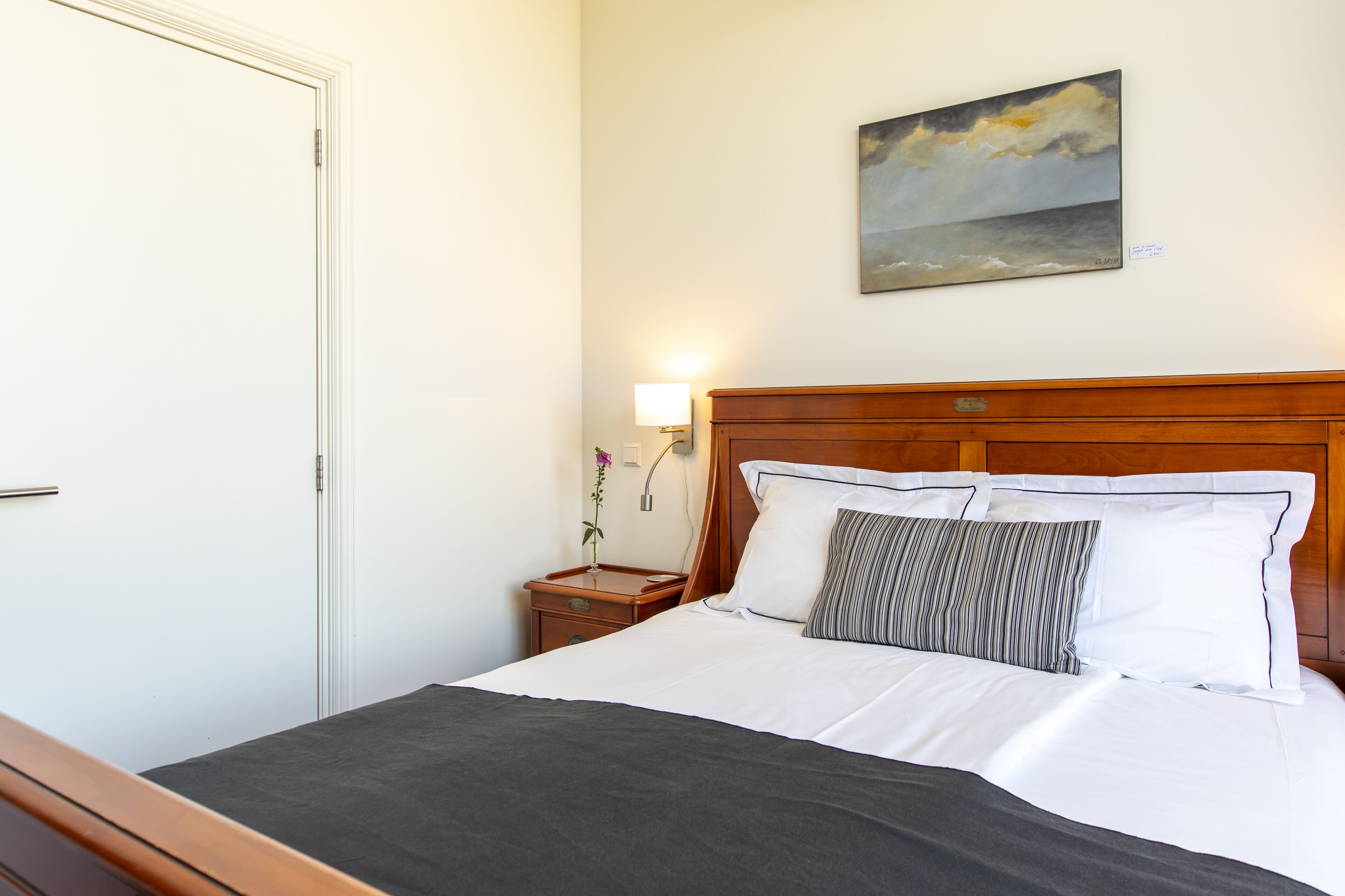 A neatly made bed with white linens and a dark gray blanket, a wooden headboard, a nightstand with a lamp, and a painting of a seascape on the wall.