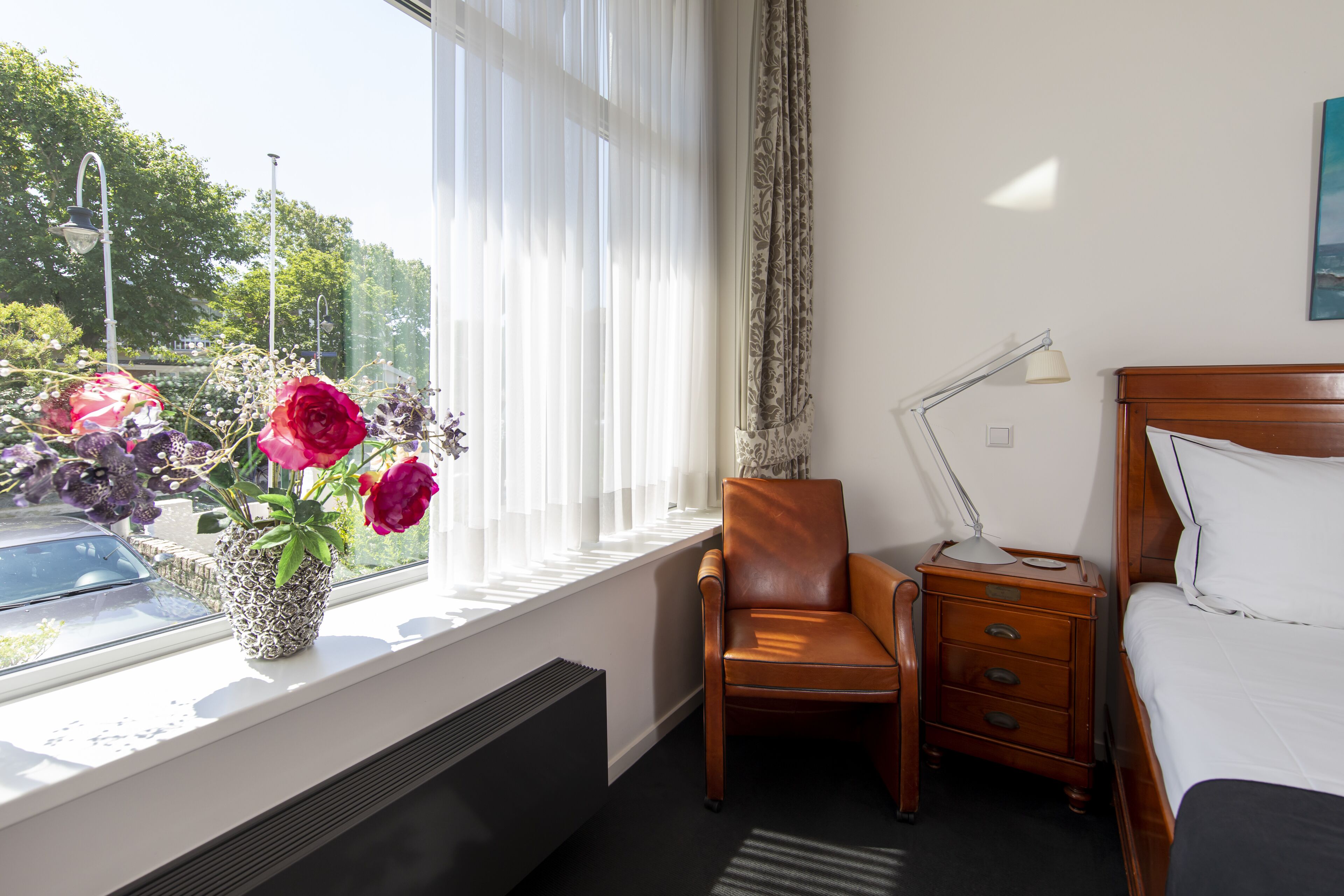 Habitación de hotel con cama, una silla, una mesita de noche, lámpara y ventana con vistas a árboles y un coche.