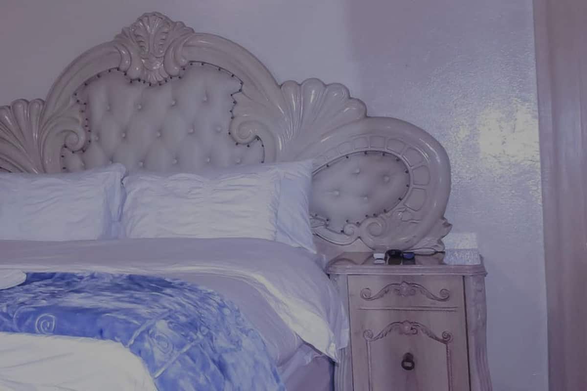 Una cama con cabecera blanca y colcha azul, una mesita de noche con cajón, y una puerta con una cruz.